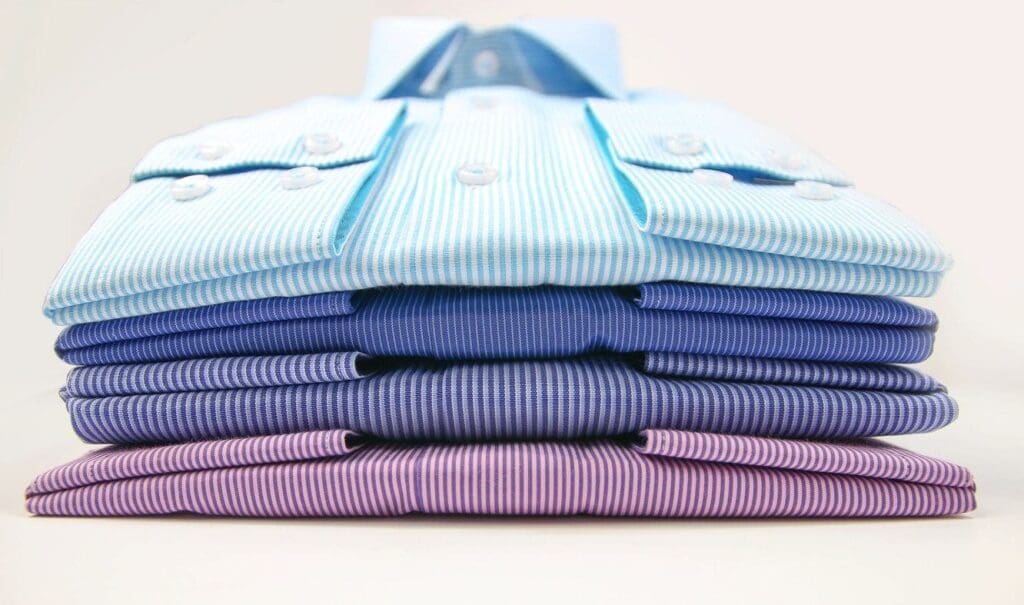 Five neatly folded dress shirts in shades of blue and purple, stacked on top of each other, viewed from the front.