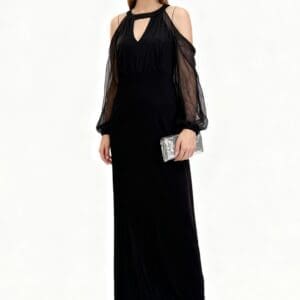 A woman wearing a long black dress with sheer sleeves, silver earrings, metallic pointed heels, and holding a silver clutch stands against a plain white background.