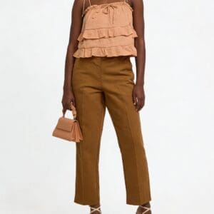 A woman stands wearing a sleeveless ruffled beige top, brown pants, beige strappy heels, and holding a small beige handbag against a plain white background.