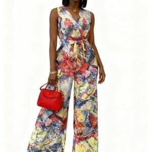 Woman wearing a colorful sleeveless jumpsuit with wide-leg pants, holding a red handbag and accessorized with gold jewelry, standing against a white background.