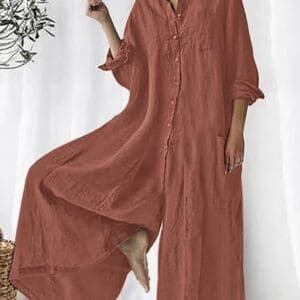 A person wearing a loose, rust-colored Cotton Linen Wide Leg Button Down Jumpsuit (SIZE L) stands indoors, lifting one foot slightly. A leafy plant appears on the left side.