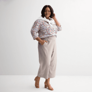A woman stands against a plain background wearing a floral top, light-colored wide-leg pants, tan heels, and a white sweater draped over her shoulders.