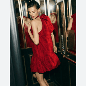 A woman in a textured red sleeveless dress stands in a mirrored elevator, looking over her shoulder, with one hand near the control panel.