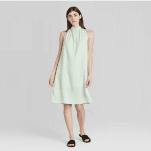A woman wearing the Tie Back Neck Dress with Pockets in size L, a light green sleeveless knee-length dress, stands against a plain background, paired with black slide sandals.