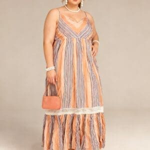 A woman wearing a peach and blue striped maxi dress, holding a small pink handbag, and standing in wedge sandals against a plain background.