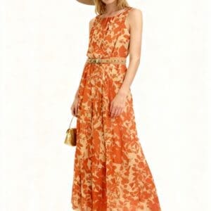 Woman wearing a sleeveless orange floral maxi dress, wide-brimmed hat, beige heels, and holding a small gold handbag, standing against a plain background.