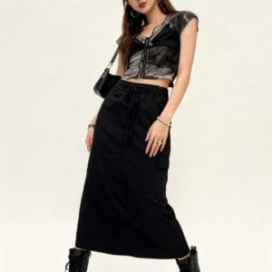 Person wearing a black cap, tie-dye cropped top, black cargo skirt, and black boots, standing against a plain light background with a casual pose.