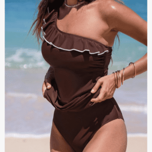 A woman wearing a brown one-shoulder swimsuit with a white ruffle stands on a beach with the ocean in the background.