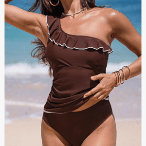 Woman wearing a brown one-shoulder bikini with white trim stands on a sandy beach with waves in the background.