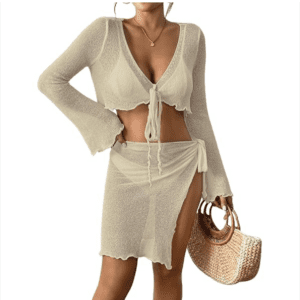 Woman wearing a sheer, light beige two-piece outfit with a tie-front crop top and wrap skirt, holding a woven straw bag. White background.