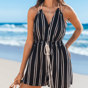 A woman wearing a black and white vertical striped romper with a rope belt stands on a sandy beach near the ocean.