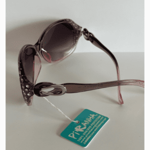 Pink sunglasses with rhinestone accents and a decorative side detail, labeled with a turquoise tag that reads "Piranha 100% UV Main Protection.