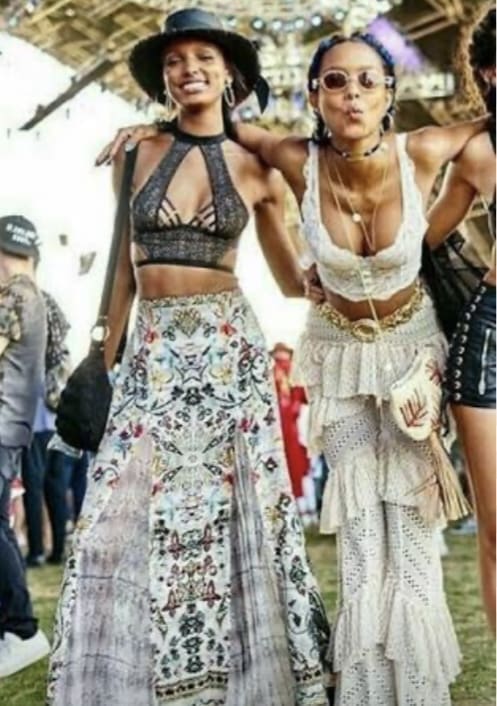 Two women at an outdoor festival wear patterned skirts and crop tops, posing and smiling for the photo. Other festival attendees and a stage are seen in the background.