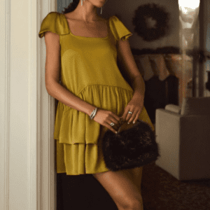 A person wearing a short, yellow tiered dress and holding a black fur clutch stands indoors near a white doorway.
