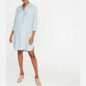 A person wearing a light blue, long-sleeve, button-up shirt dress with a chest pocket, and beige backless shoes, standing against a plain white background.
