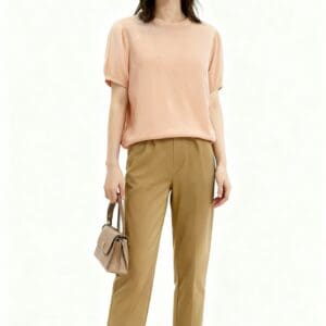 A woman wearing sunglasses, a light pink blouse, tan cropped pants, and loafers stands holding a small beige handbag against a plain white background.
