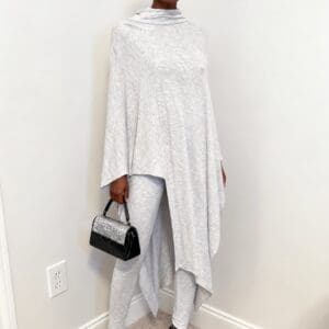 A person stands indoors wearing a gray asymmetrical poncho, matching leggings, black beret, sunglasses, silver heels, and holding a small black and silver handbag.