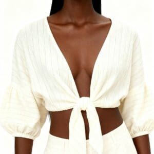 A woman wearing a white, deep V-neck, tie-front crop top with three-quarter puff sleeves and white high-waisted pants, posing against a plain background.