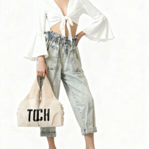 A person wearing a white tie-front top, high-waisted light blue jeans, black heeled sandals, and gold jewelry holds a beige tote bag with "TCH" text.