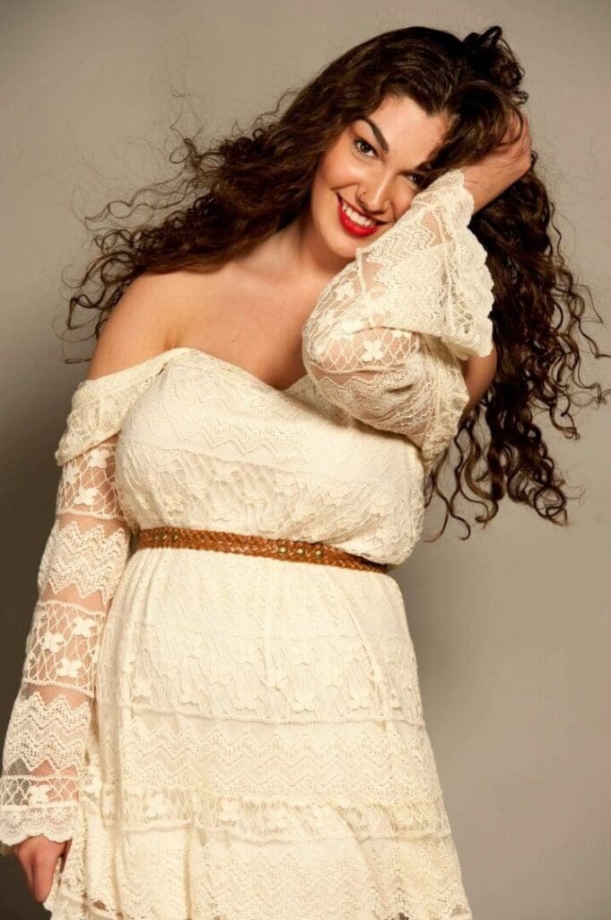 Woman with long curly hair wearing an off-shoulder white lace dress and brown belt, smiling and touching her hair against a neutral background.