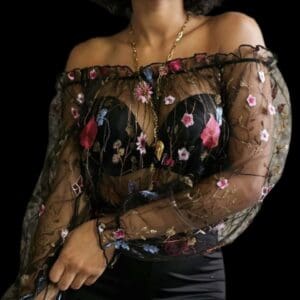 A person wearing an off-the-shoulder black sheer top with colorful floral embroidery and a long gold necklace, set against a black background.