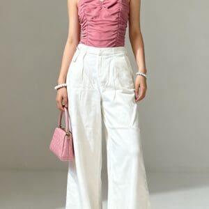 A woman stands indoors wearing a pink sleeveless ruched top, white high-waisted wide-leg pants, pink handbag, and statement earrings.