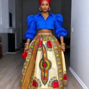 A woman stands indoors wearing a bright blue puff-sleeve crop top, a red headwrap, and a long, patterned skirt with colorful African prints. She holds a red clutch and wears gold jewelry.