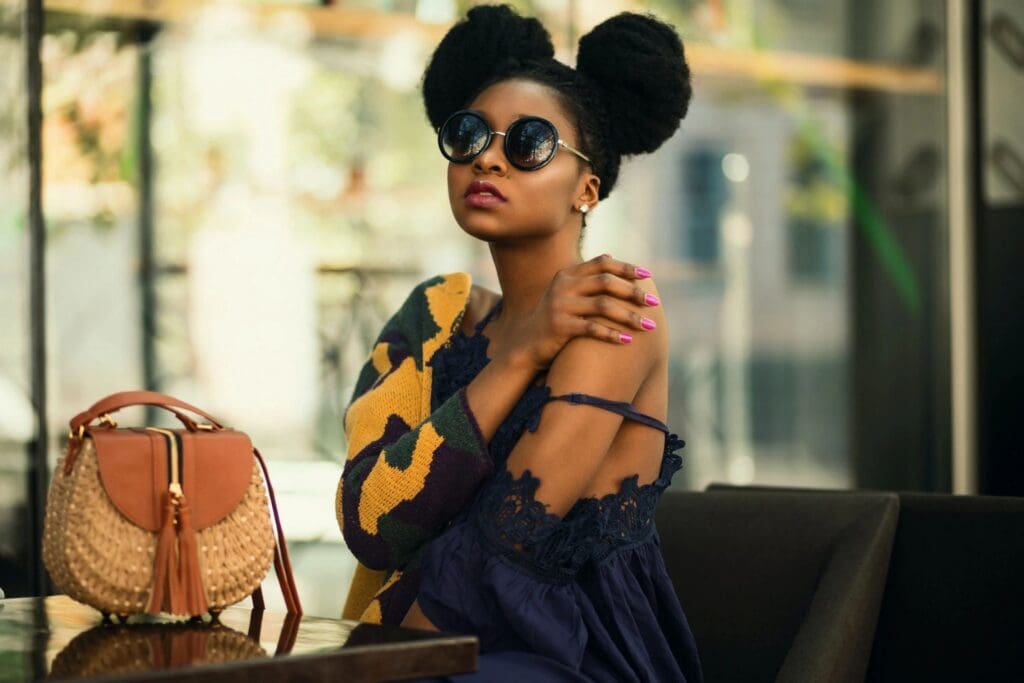 Woman with natural hair in two puffs wearing sunglasses, a dark blue dress, and a patterned sweater sits at a table with a woven handbag beside her.