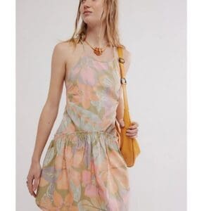 Woman wearing a pastel floral sleeveless dress, carrying a yellow shoulder bag, and accessorizing with a chunky necklace, stands against a plain light background.