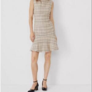 A woman stands against a plain background, wearing a sleeveless, knee-length plaid dress and black heeled shoes.