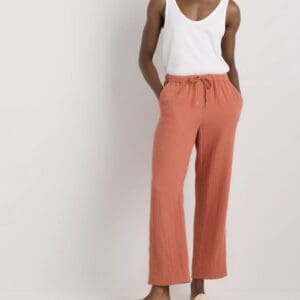 Person wearing a white sleeveless top, rust-colored wide-leg pants with a drawstring waist, and beige sandals, standing against a neutral background.