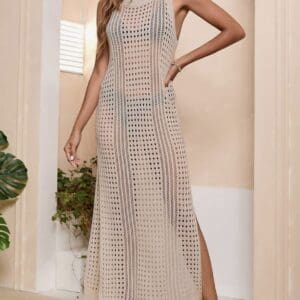 Woman wearing a sleeveless, beige crochet maxi dress with side slits, standing barefoot indoors against a light-colored wall with plants.