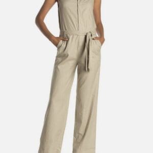Woman wearing a sleeveless, beige belted jumpsuit with wide-leg pants and V-neckline, standing with hands in pockets and wearing nude heels.