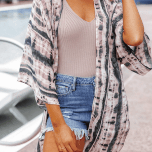 Woman wearing a tie-dye kimono over a beige ribbed tank top and denim shorts, standing outdoors by a pool.
