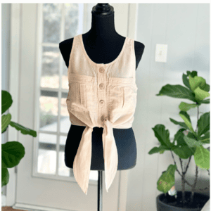 Sleeveless beige button-front crop top with tie waist displayed on a black dress form. Background includes indoor plants and a glass door.