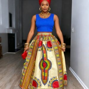 Woman standing indoors wearing a red headwrap, blue sleeveless top, colorful patterned maxi skirt, and gold jewelry, holding a red clutch.