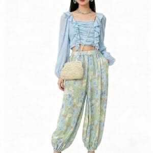 Woman wearing a light blue ruffled blouse, patterned loose pants, heeled sandals, hoop earrings, a necklace, and a woven crossbody bag, standing against a plain white background.