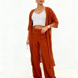 Woman stands in front of a white background wearing a rust-colored outfit, white crop top, white sneakers, gold jewelry, and holding a small white purse.