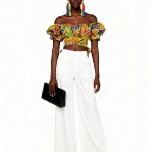 Woman standing against a white background, wearing a colorful off-shoulder crop top, white wide-leg pants, black heels, rainbow tassel earrings, and holding a black clutch.