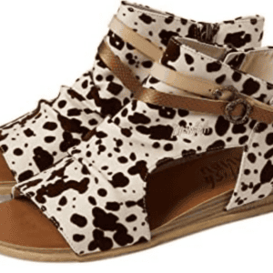 A pair of open-toe flat sandals with a white and brown cow print pattern, side zipper, and thin tan decorative strap.