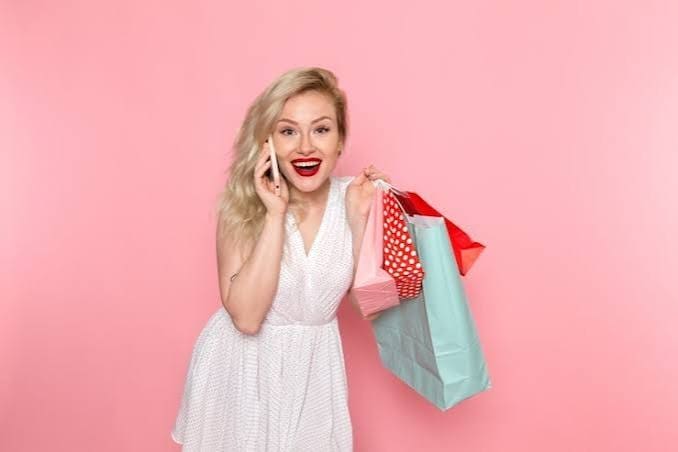 A woman in a white dress smiles while holding shopping bags and talking on a phone against a pink background.