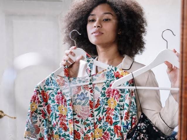 A person stands in front of a mirror holding up two floral print dresses on hangers, appearing to decide which one to wear.