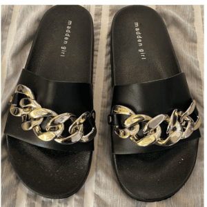 A pair of black Madden Girl slide sandals with large gold chain accents on each strap, placed on a striped fabric background.