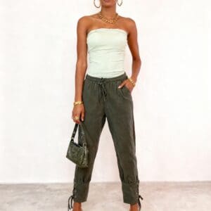 A woman stands against a plain wall wearing a strapless top, olive pants, sandals, sunglasses, and gold jewelry, holding a small handbag.