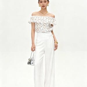 Woman stands facing forward, wearing a white off-shoulder top with floral print, wide-leg white pants, sandals, gold jewelry, and holding a small silver handbag against a plain white background.