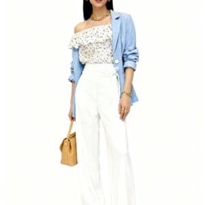 Woman wearing a blue blazer, floral off-shoulder top, white high-waisted pants, brown sandals, and gold jewelry while carrying a tan handbag, standing against a white background.