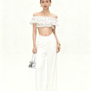 Woman stands against a plain white background wearing an off-shoulder floral crop top, white wide-leg pants, sandals, and holding a small handbag. She has her hair in a bun and wears jewelry.