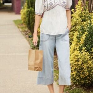 A person wearing a white embroidered top, light blue cropped jeans, and sandals holds a brown paper bag while standing on a sidewalk near green bushes.