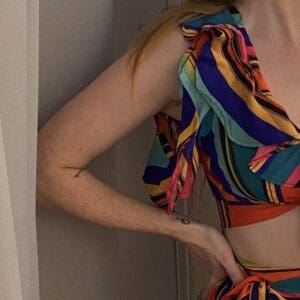Person wearing a colorful, striped, ruffled crop top and skirt stands near white curtains with one hand on their hip.