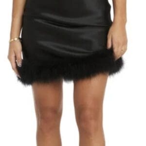 Woman wearing a sleeveless black satin mini dress with a feather trim at the hem, clear heeled sandals, and minimal jewelry.